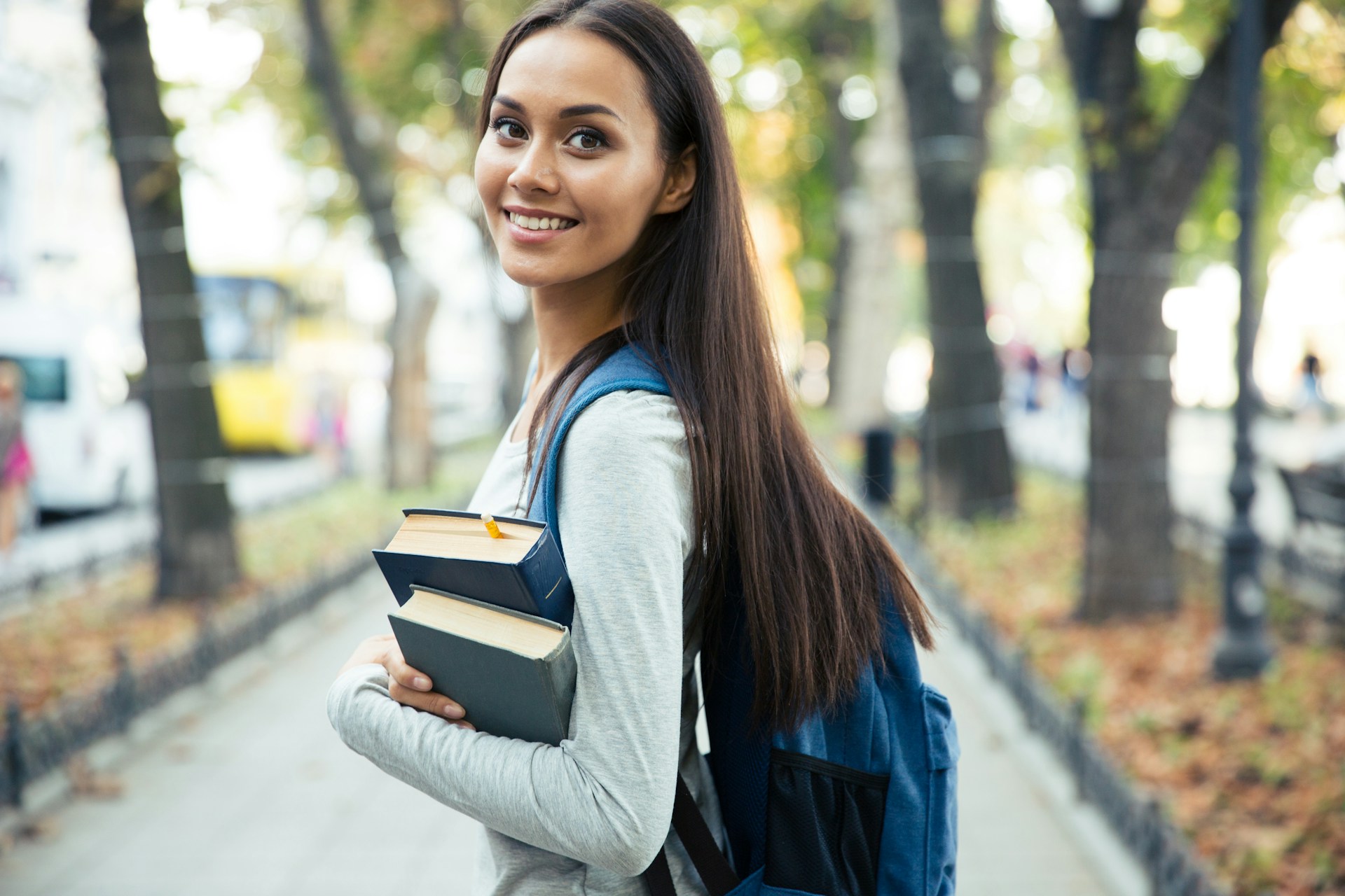 A young, female university student carries two hardbound books in her arms while wearing a backpack and walking along a blurred path lined with trees, representing child support for adult children.